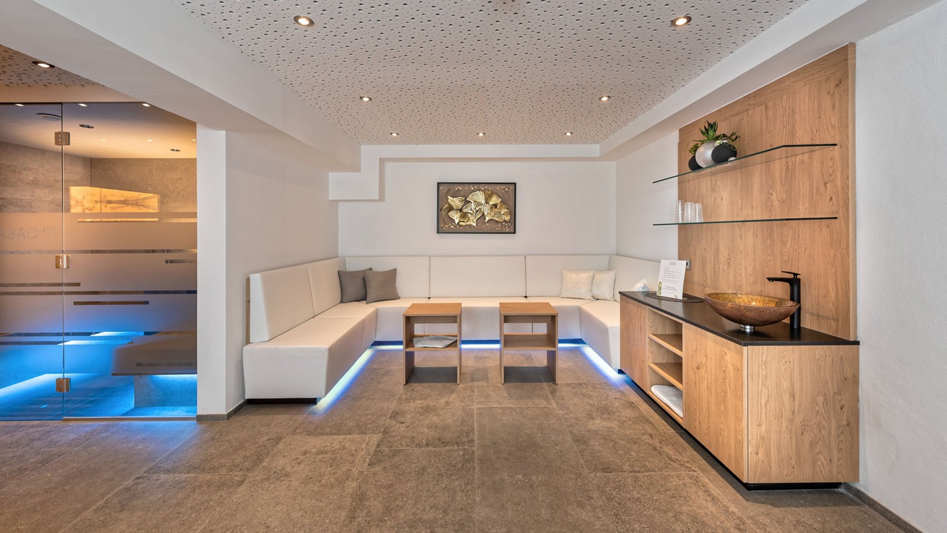 Stubaital: family hotel for holidays in comfort Modern wellness waiting area with white sofa and wooden furniture next to a blue-lit room